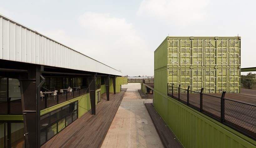 Green shipping containers form a building with a walkway. Outdoor setting with a metal roof and fence.