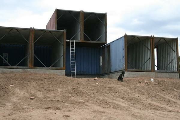 Shipping containers, stacked, on dirt mound; a dog sits nearby, ladder at container entrance.