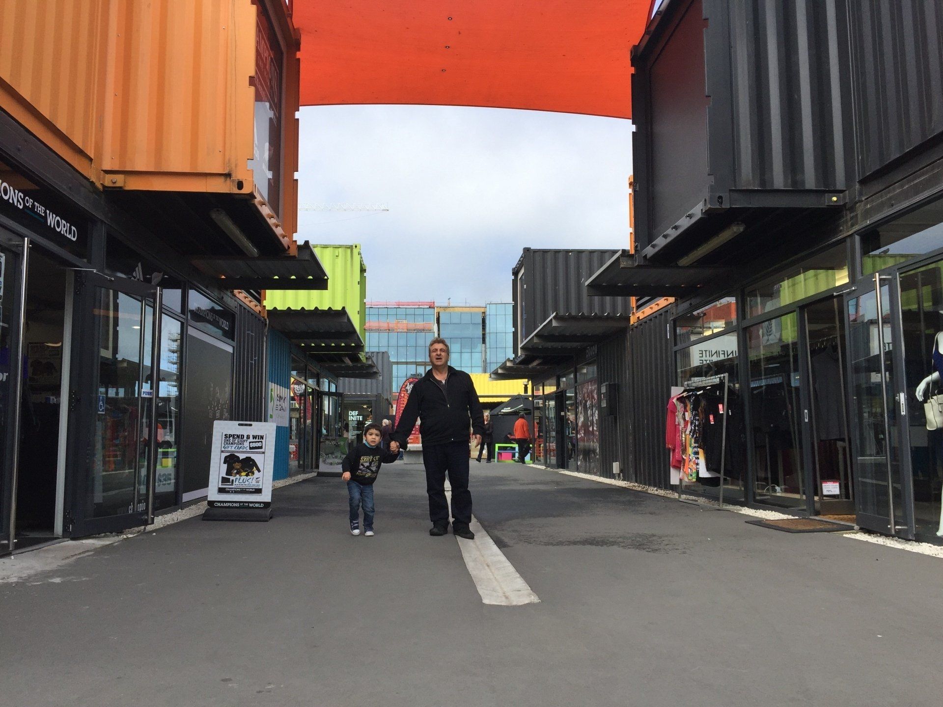 Father and child walking hand-in-hand through a shopping area made of shipping containers.