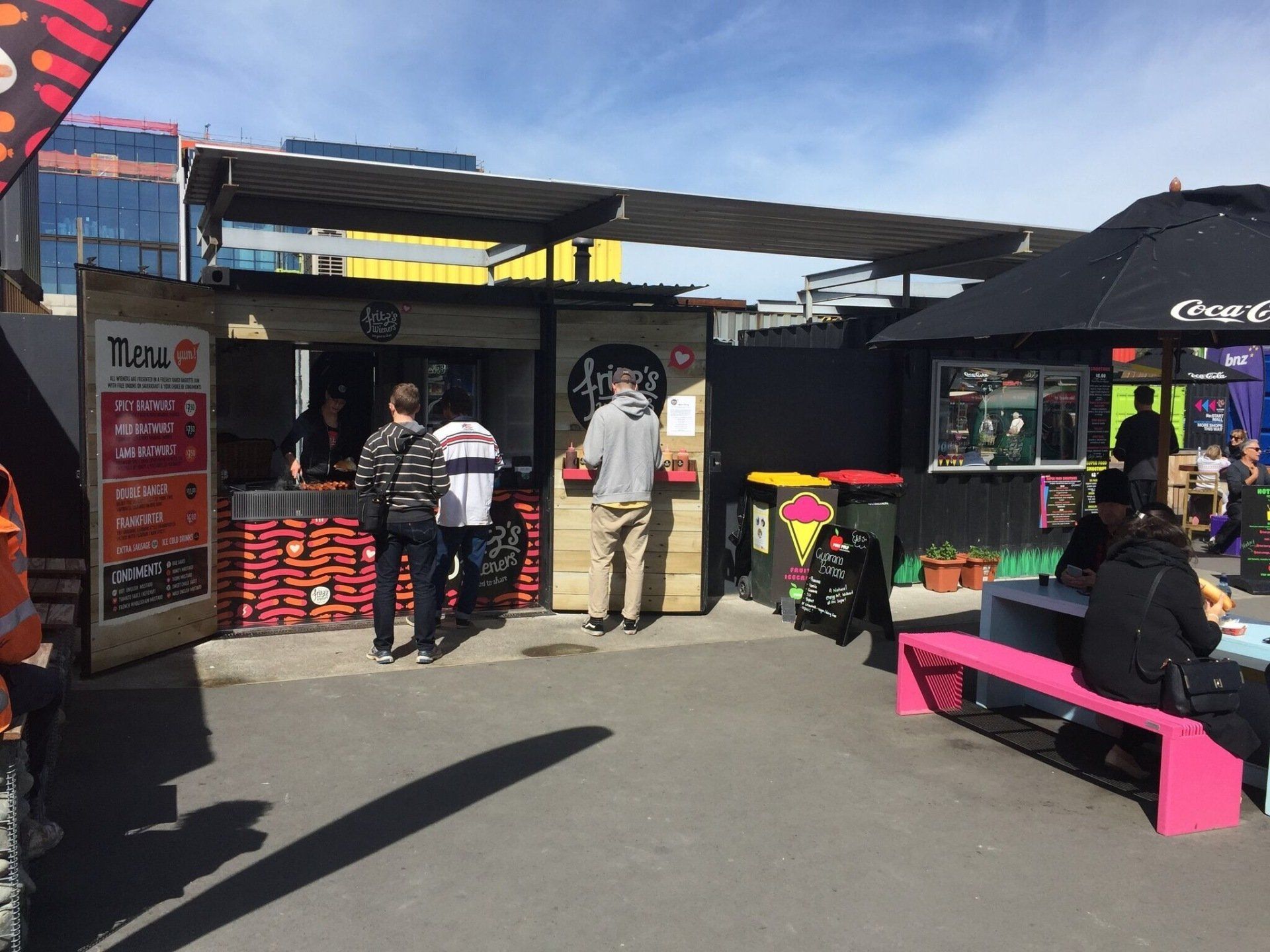 Food stalls in an outdoor market with customers. Buildings and blue sky in the background.