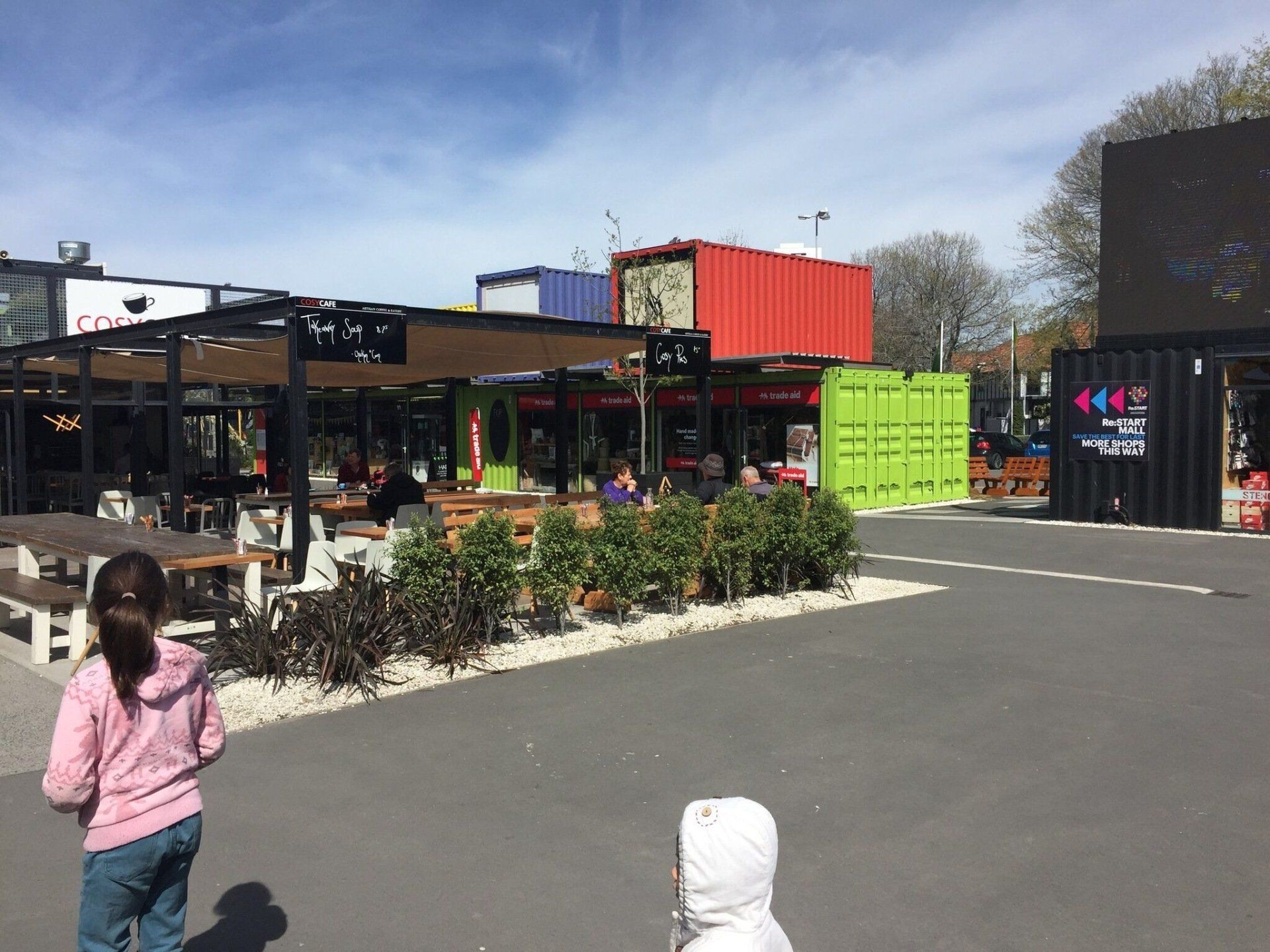 A plaza with colorful shipping containers transformed into shops and restaurants. A child walks in the foreground.