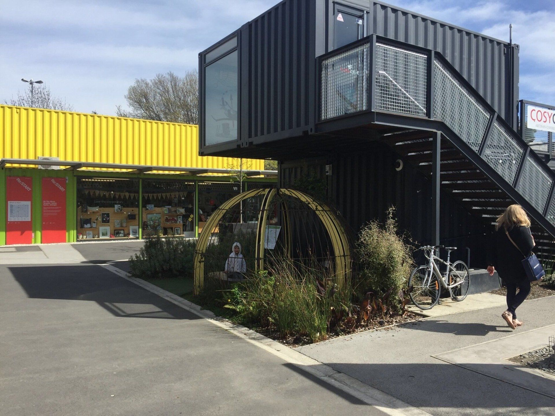 Black container building with outdoor staircase and yellow building in the background. A person walks by.
