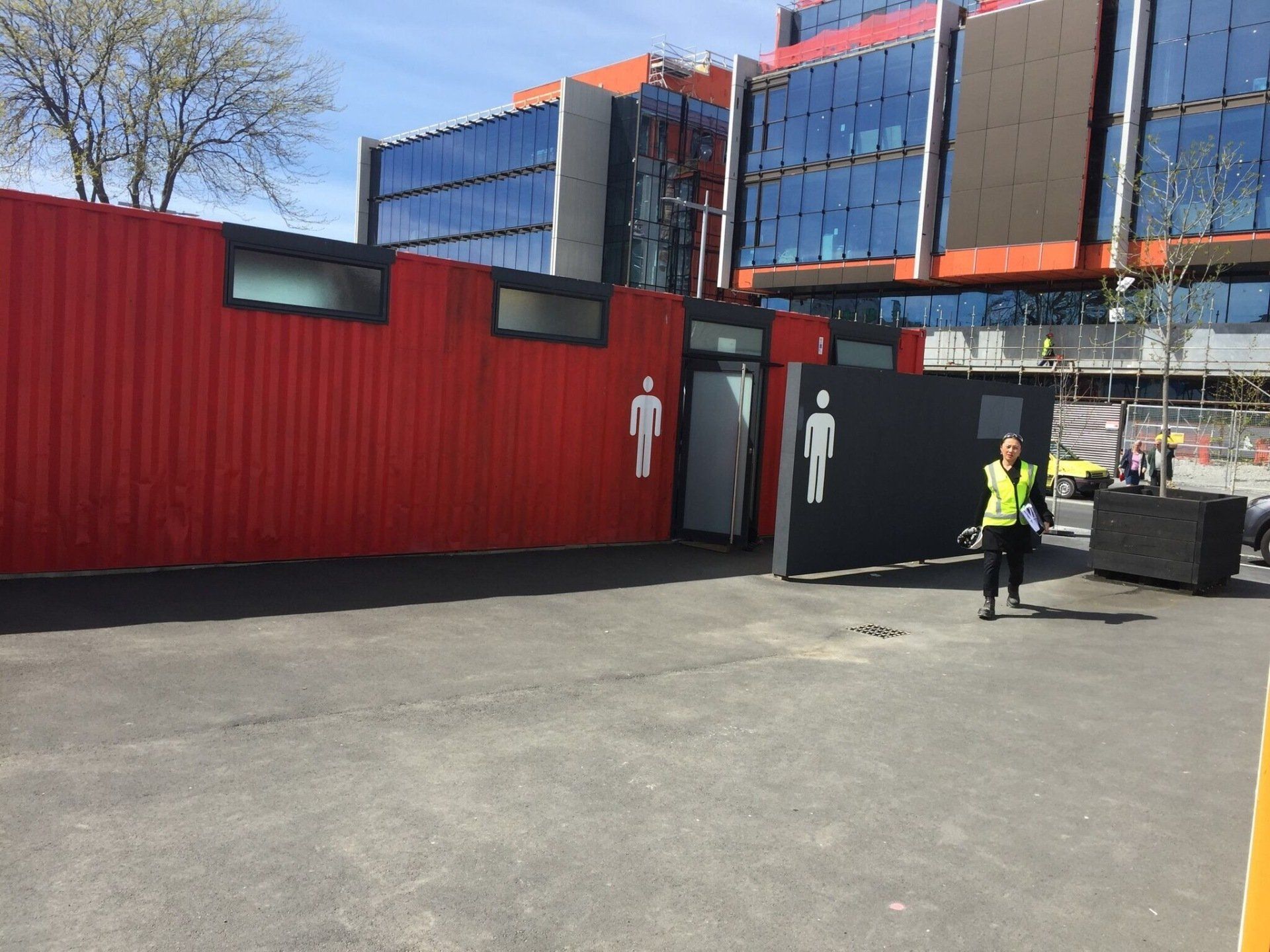 Red and black container restrooms with male symbols; a person in a vest stands near it in a parking lot.