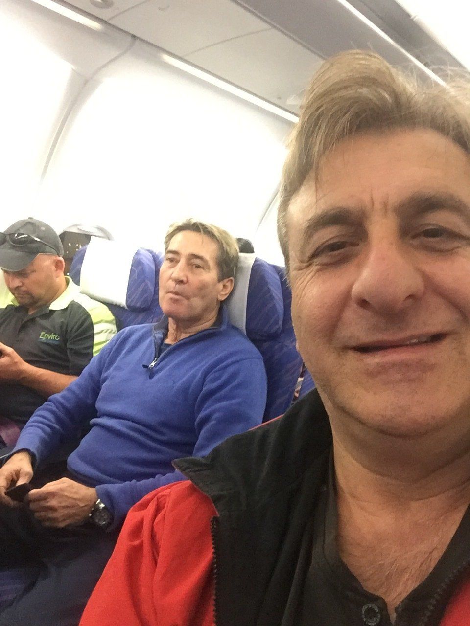 Man takes a selfie on a plane with two other passengers. One in a blue shirt, the other in a green shirt.