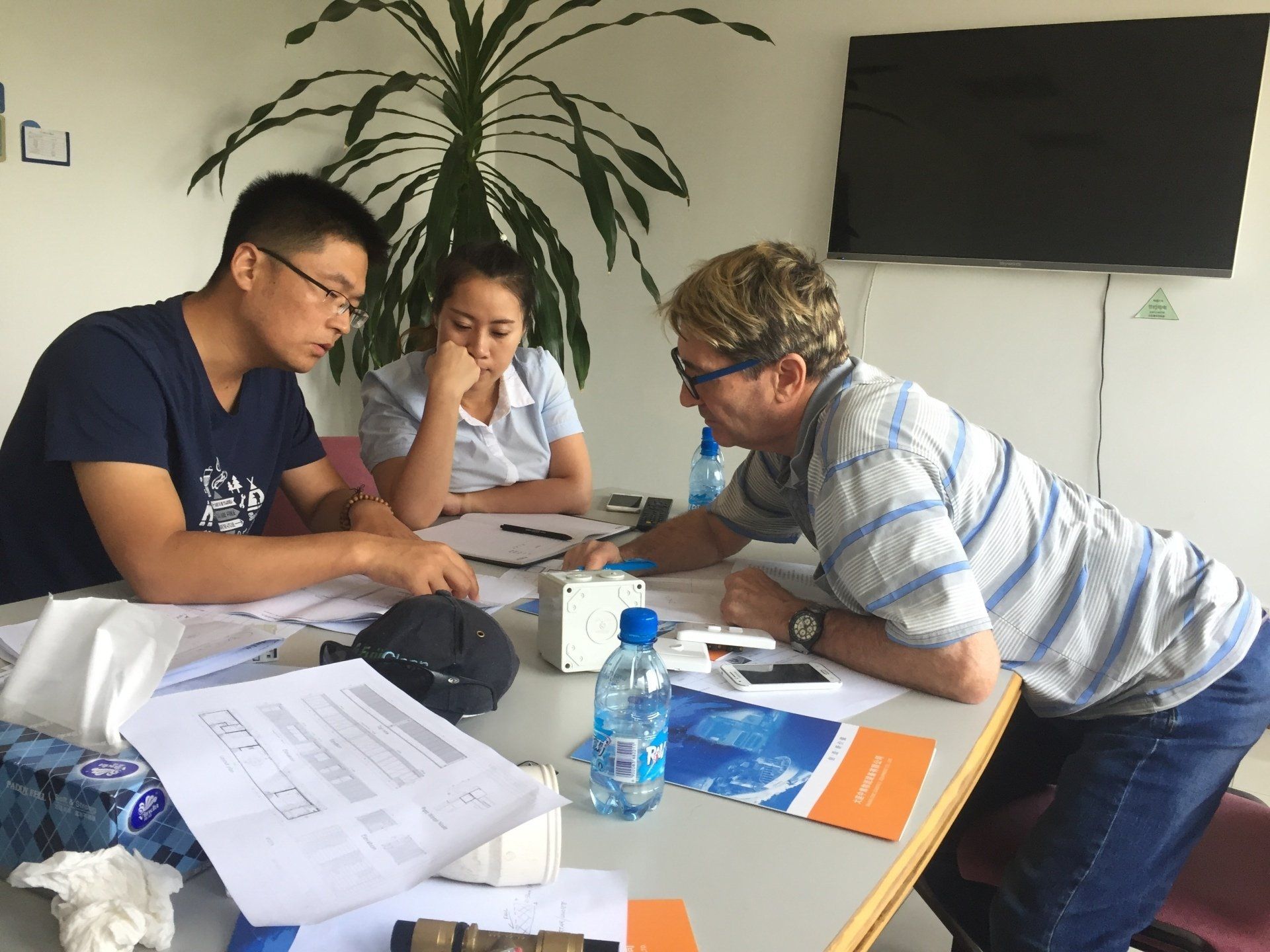 Three people reviewing papers at a table. Two Asian people and one white person lean in closely. Papers and a tissue box are on the table.