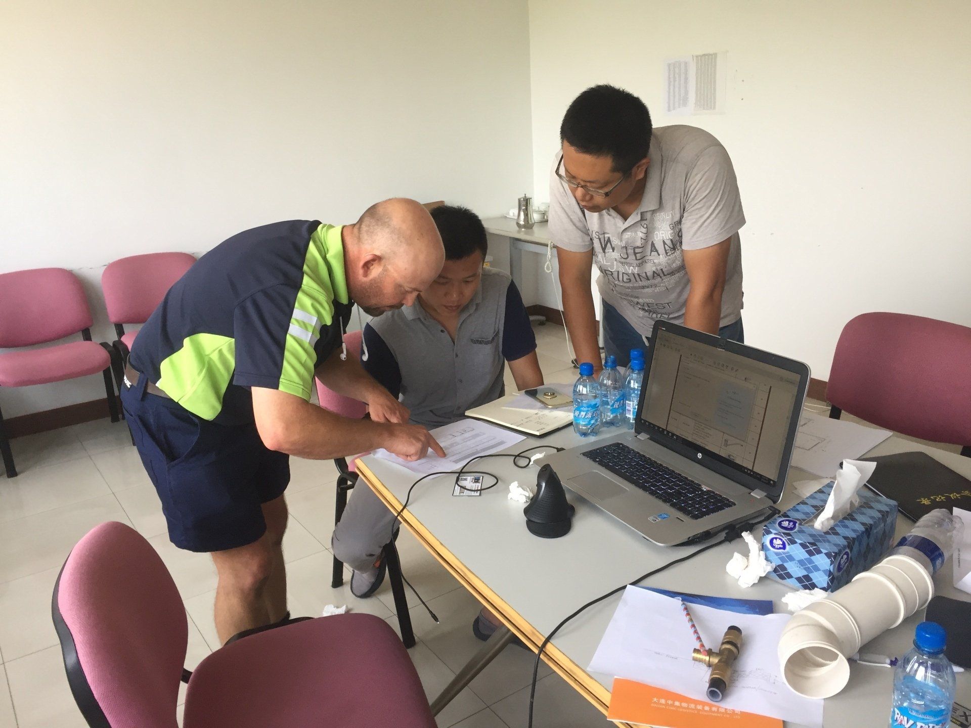 Three people examining papers and laptop at a table, possibly discussing a project; bottles of water and tools visible.