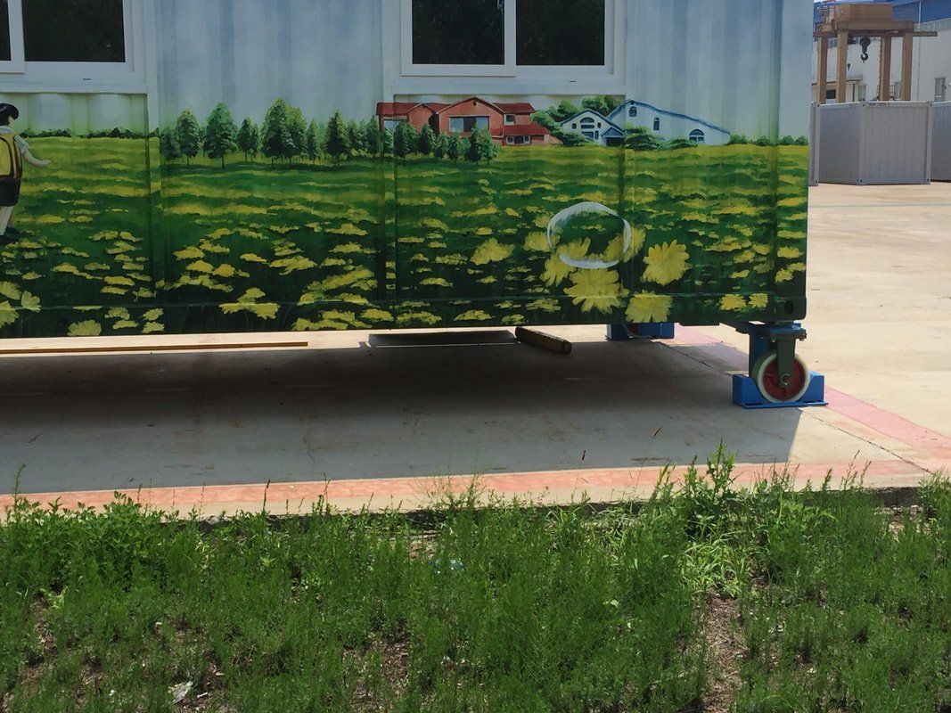 Side of a building with painted mural of a field of yellow flowers and a rural scene; on blue blocks with red wheels.