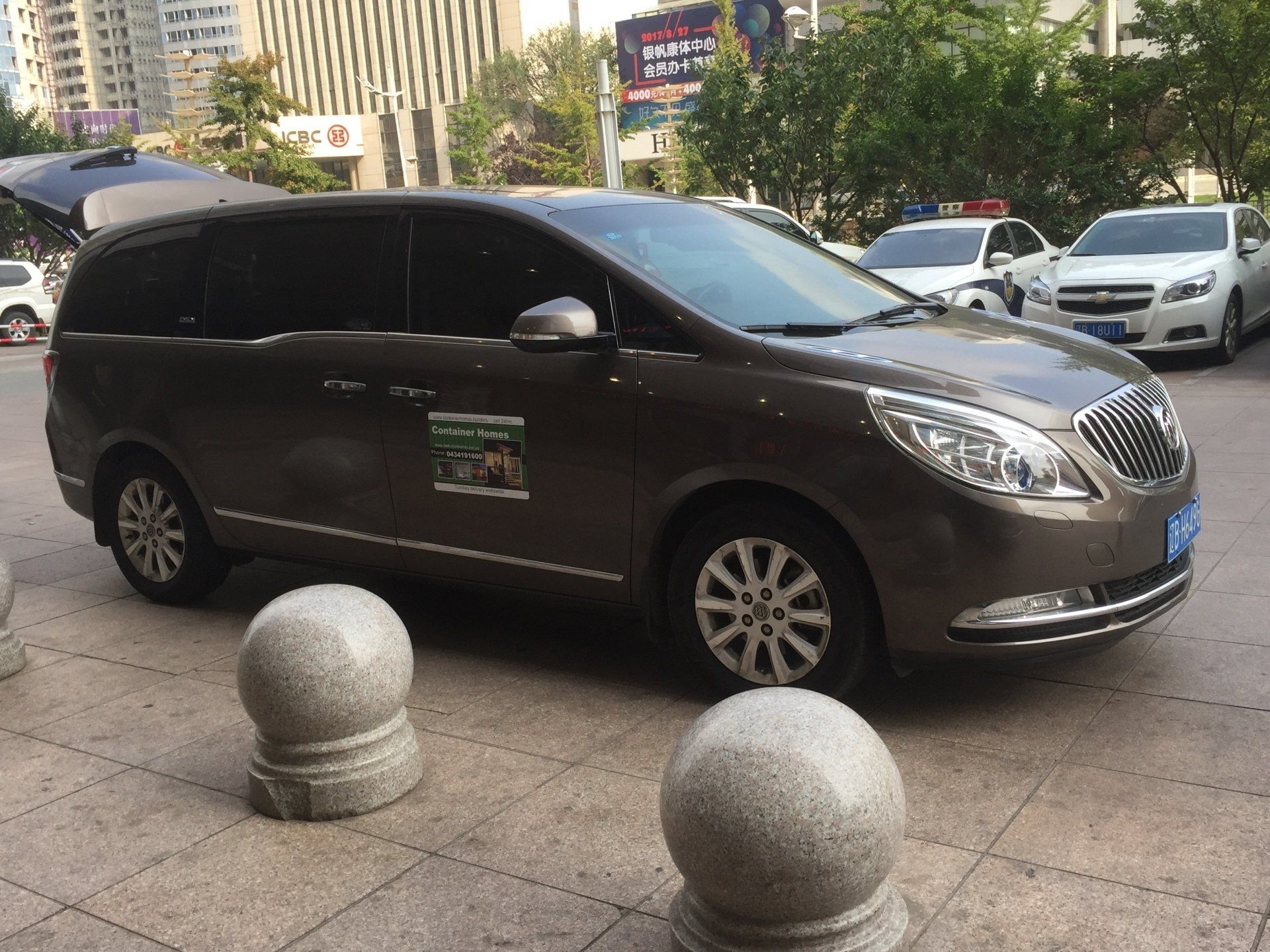 Brown minivan parked on a sidewalk next to stone bollards.  Other cars and buildings are in the background.