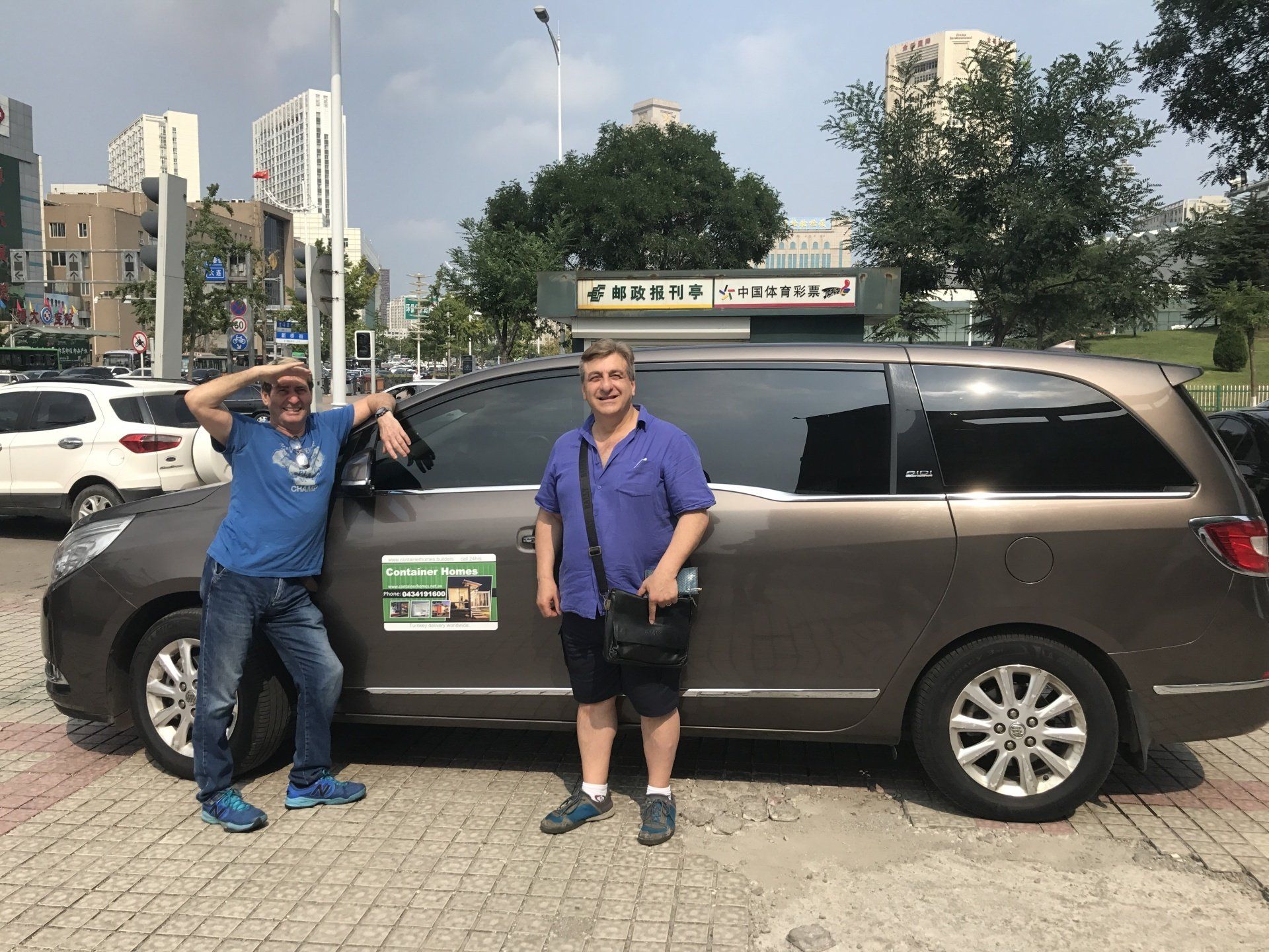 Two men pose next to a brown minivan outdoors. One man gestures towards the vehicle, the other holds a bag.