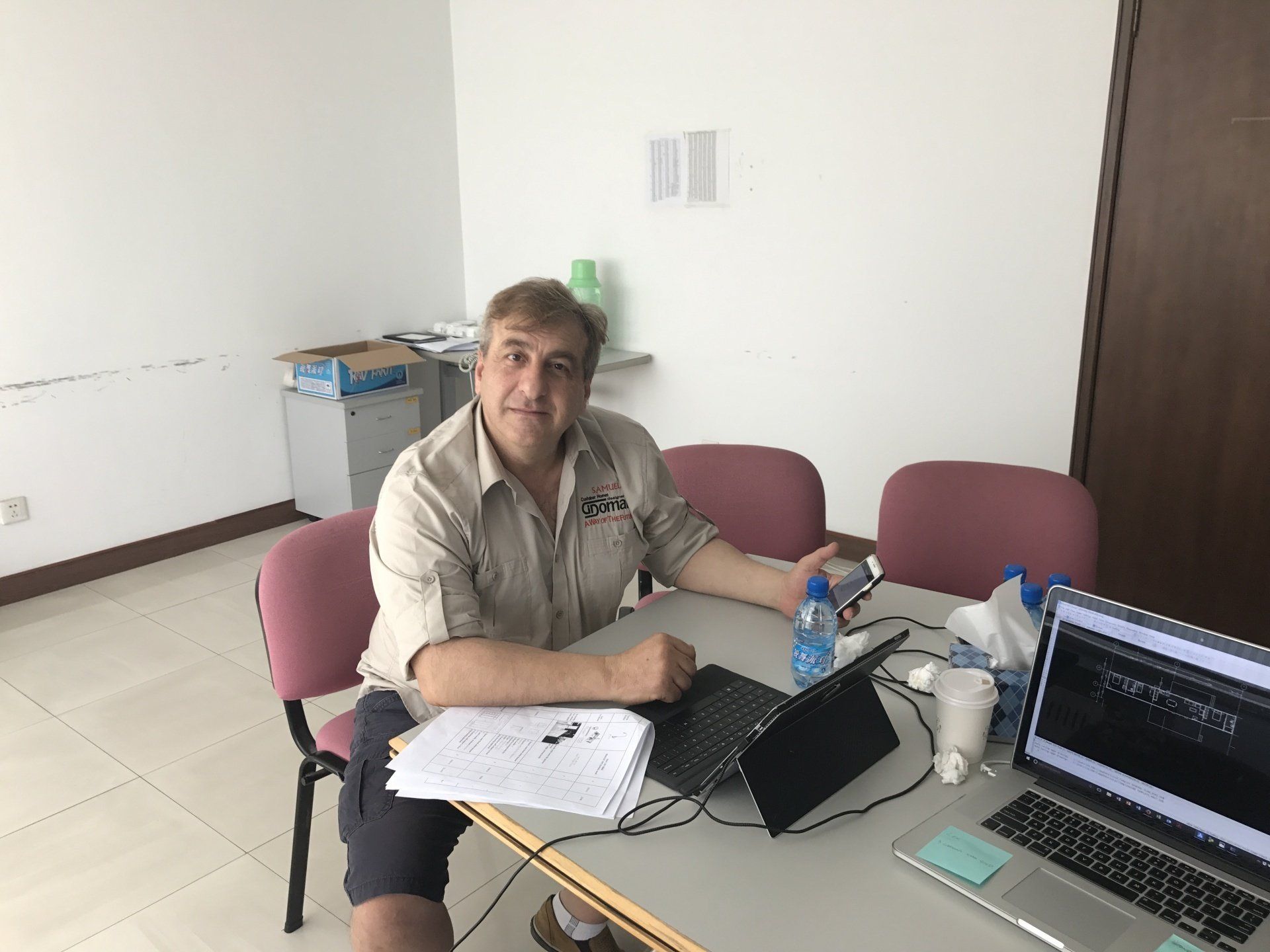 Man seated at table with laptop, papers. Wearing light brown shirt. White room.