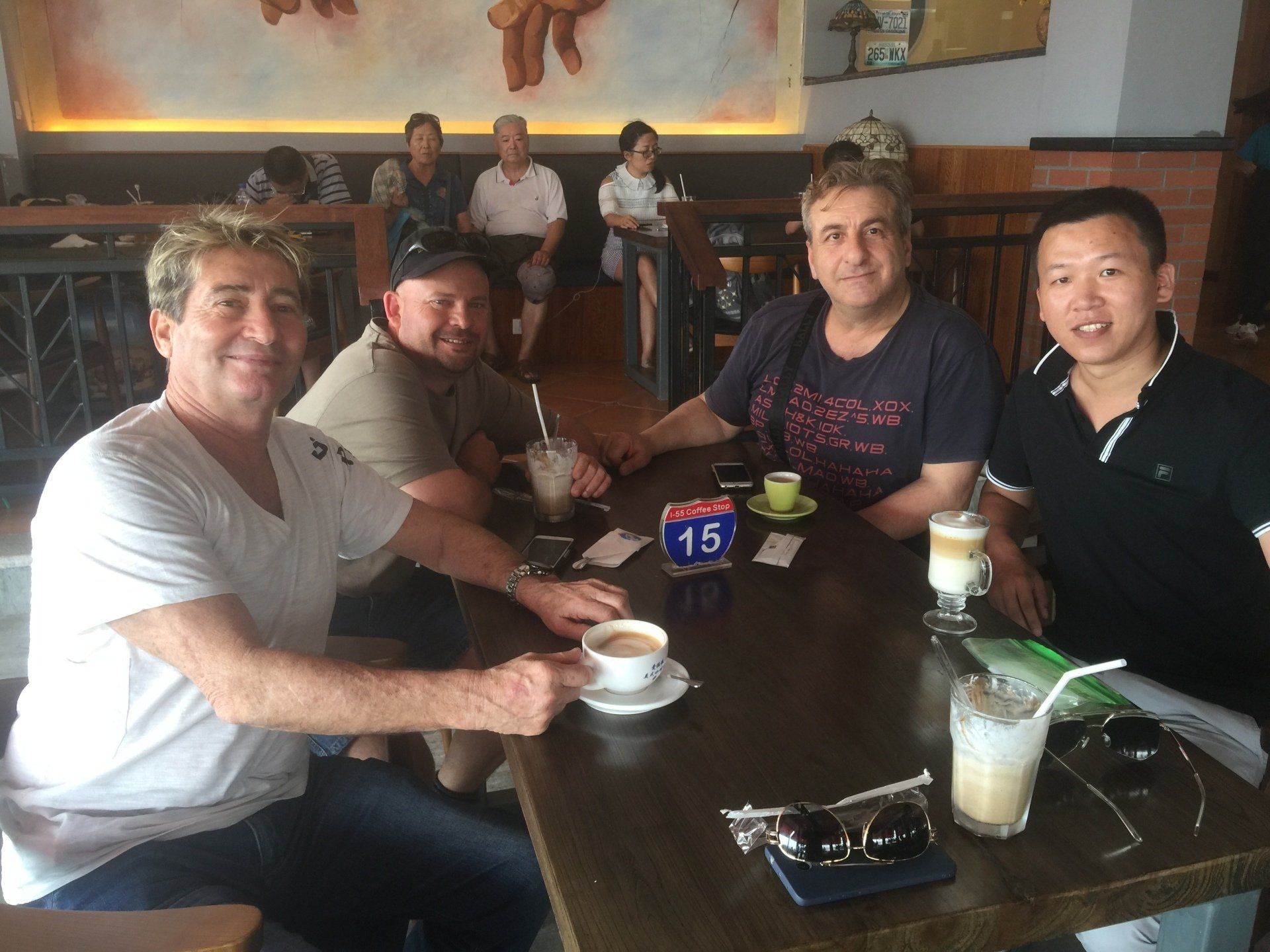 Four people at a cafe table with coffee and drinks. Others in background.