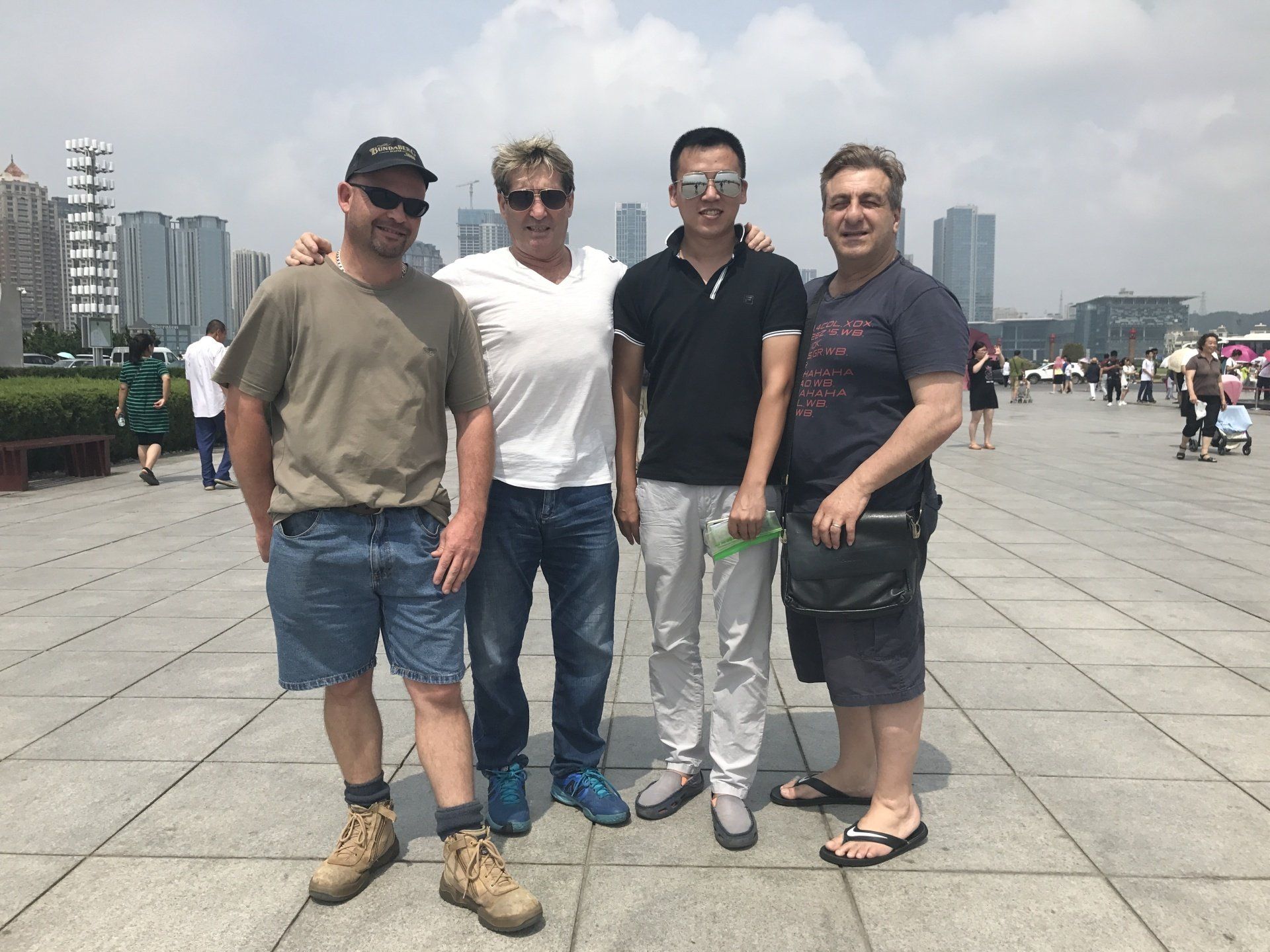 Four men standing and posing outside near buildings on a sunny day.