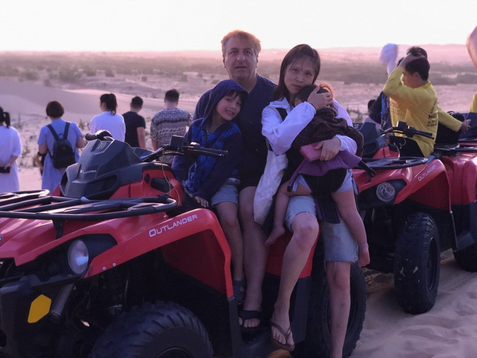 Family poses on red ATVs in a desert. A man and woman hold children, other people in background.