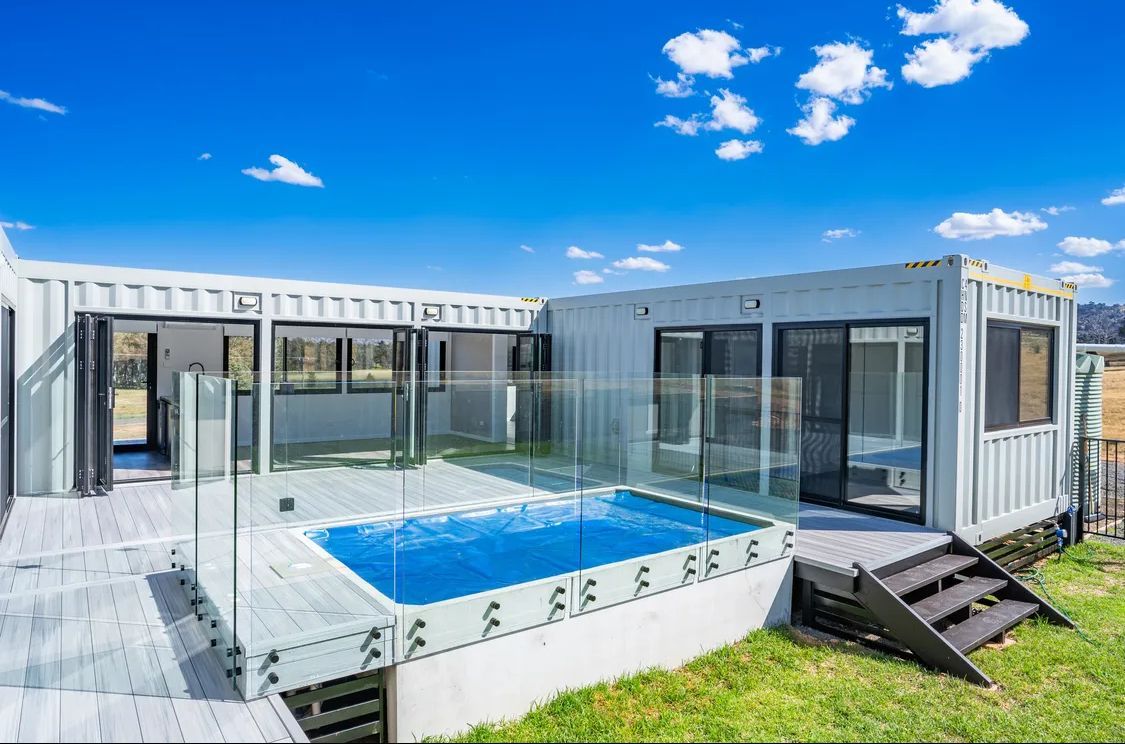Shipping container home with a small pool, surrounded by a deck and glass railing, on a grassy hill under a blue sky.
