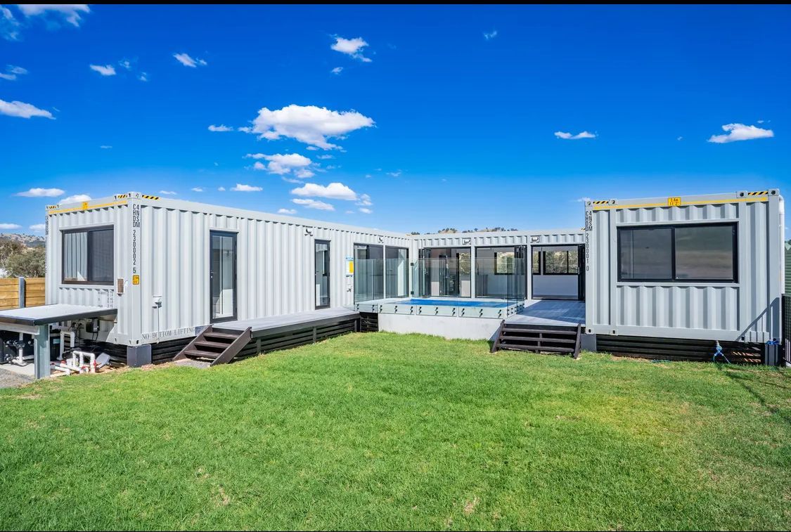 White shipping container home with deck, pool, and green lawn against a blue sky.