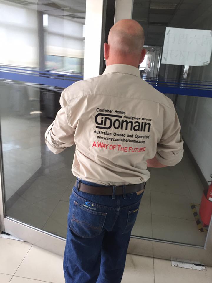 Man in beige shirt and jeans, back to the camera, standing by a glass door. Shirt has logo: 