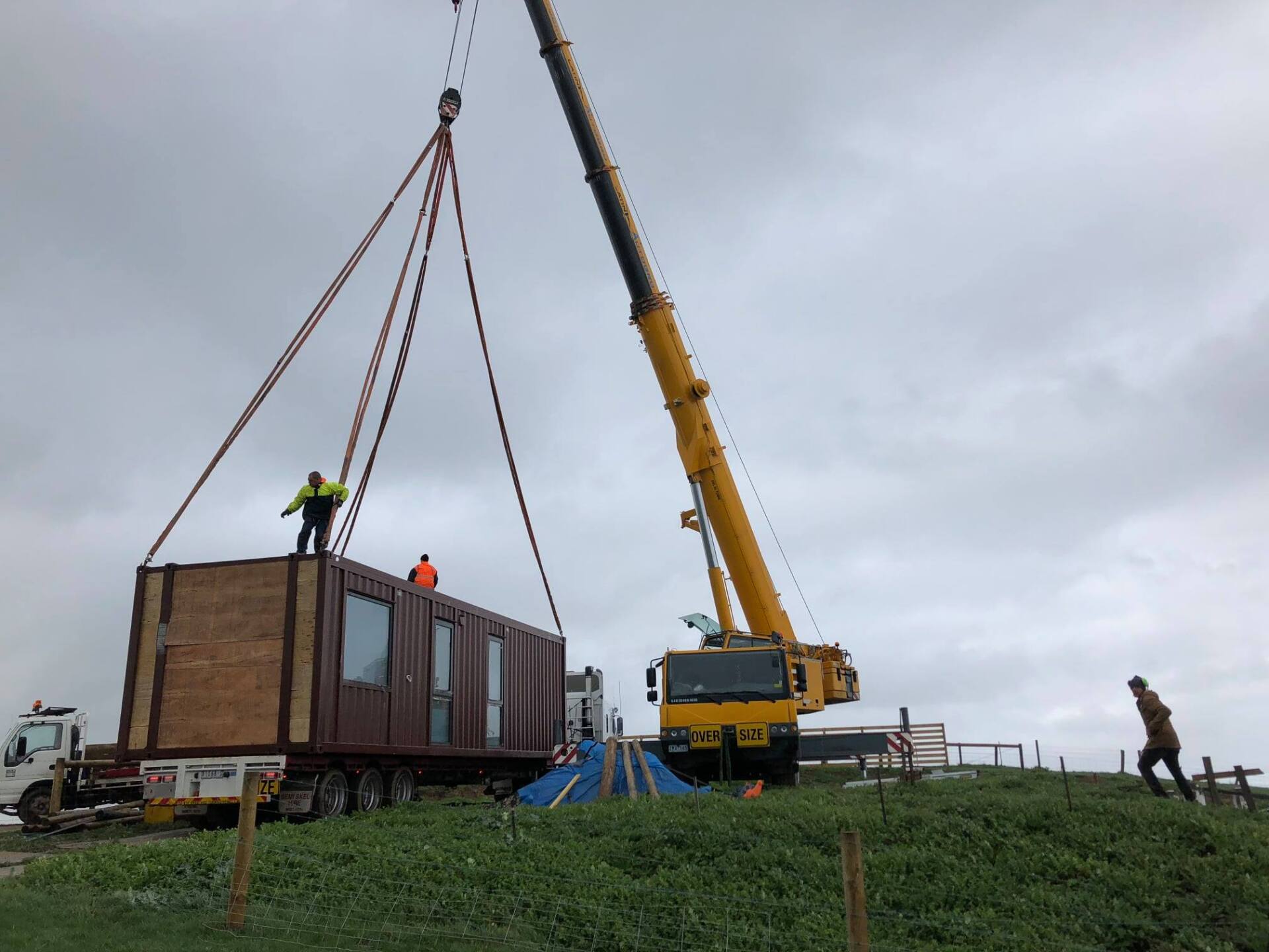 A crane lifting a modular building off a truck on a grassy hill. Workers oversee the process under a cloudy sky.