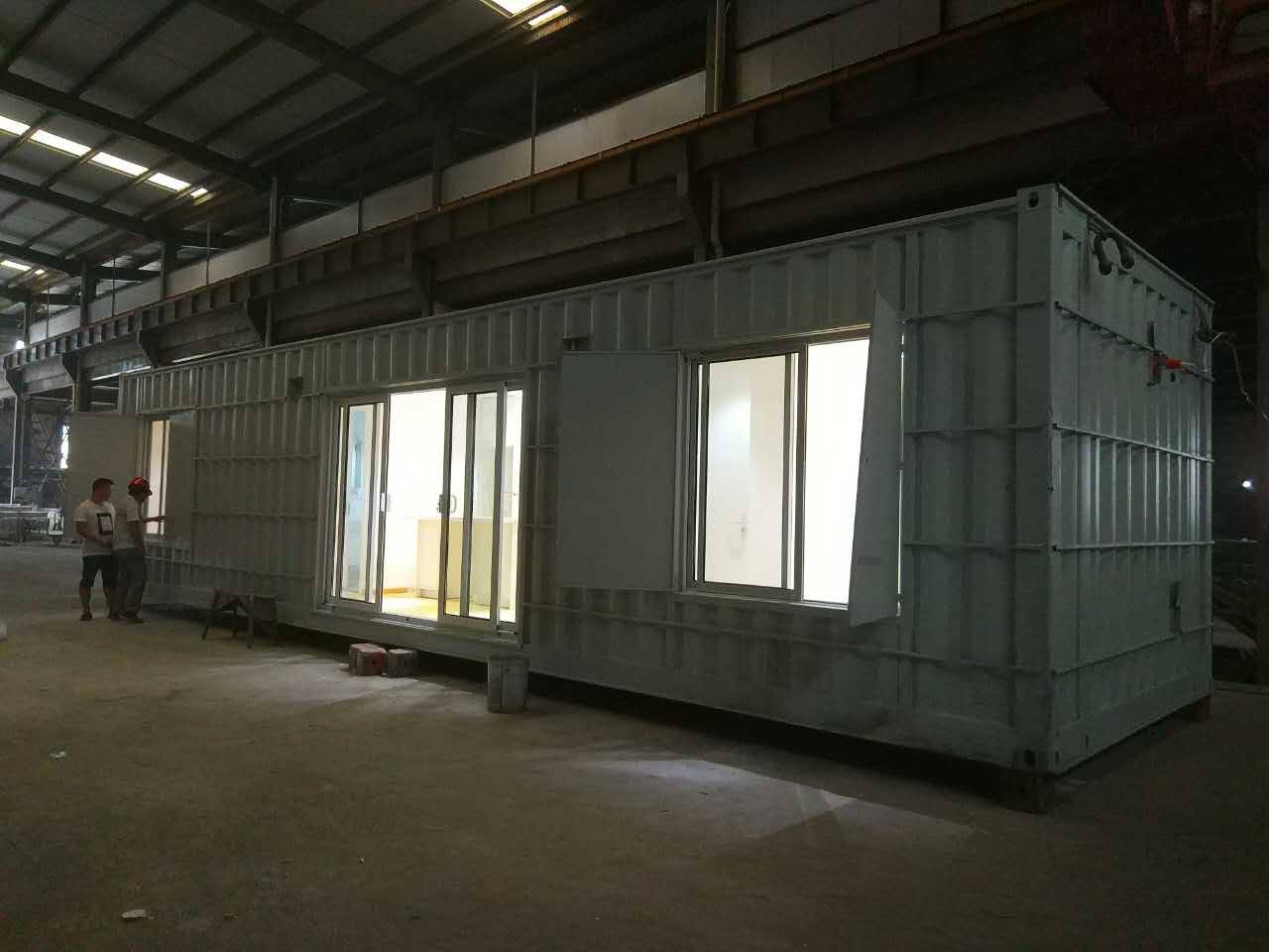 White shipping container building with open doors/windows, people inside, industrial setting.