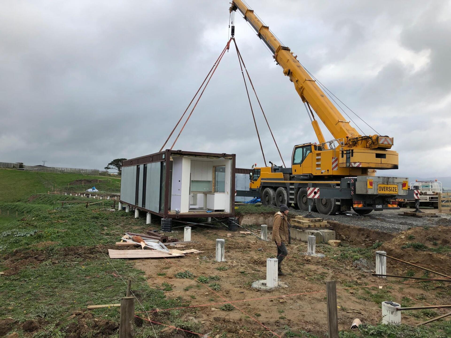 Crane lifting a container-like building onto concrete pillars at a construction site on a cloudy day.