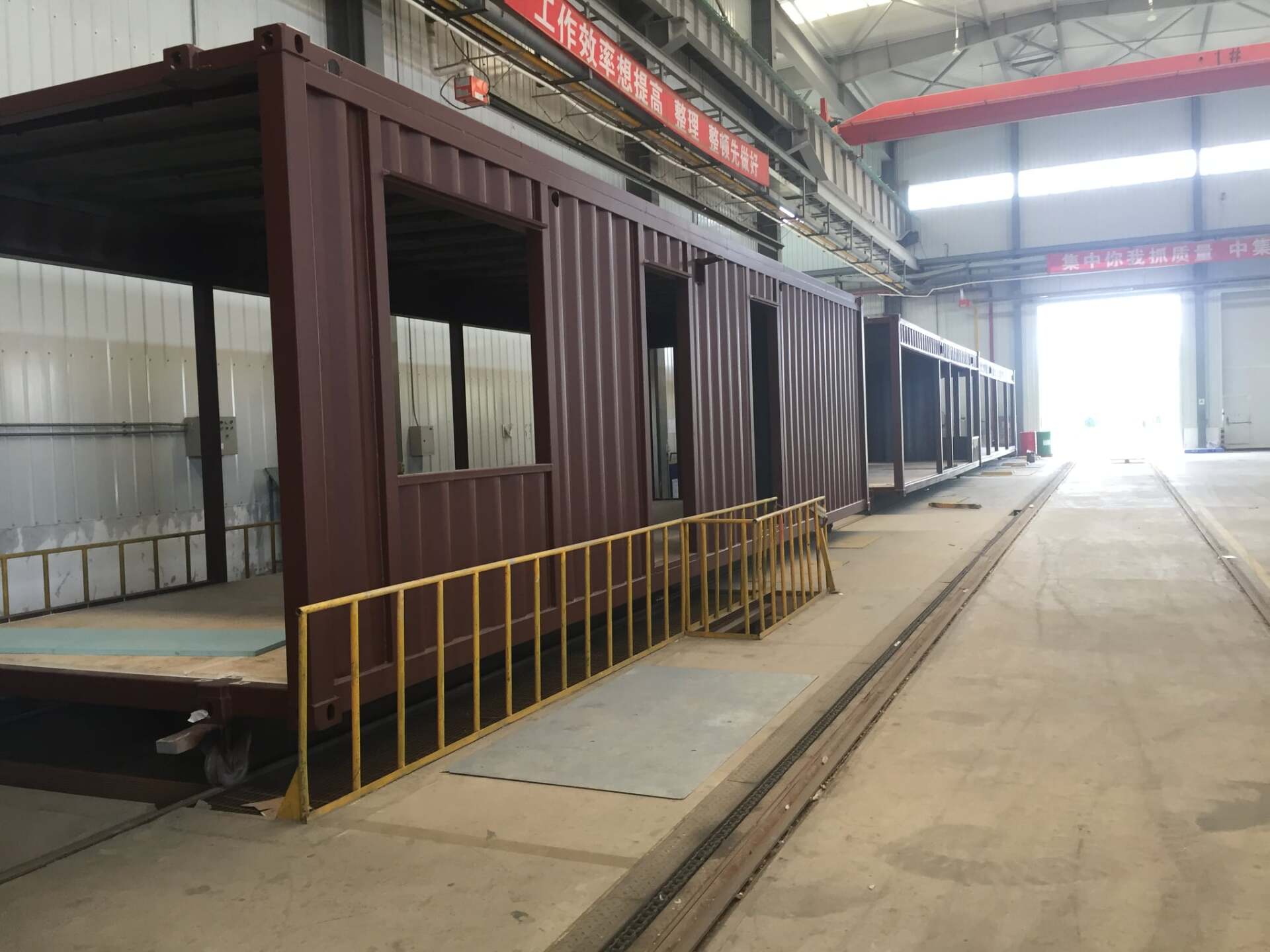 Brown container structure inside a factory. It has open sections for windows and doors and a yellow railing.
