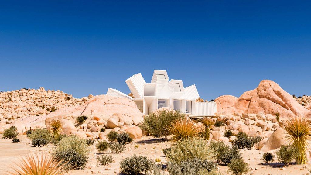White geometric house in desert landscape, surrounded by tan rocks, green and orange bushes, and a clear blue sky.