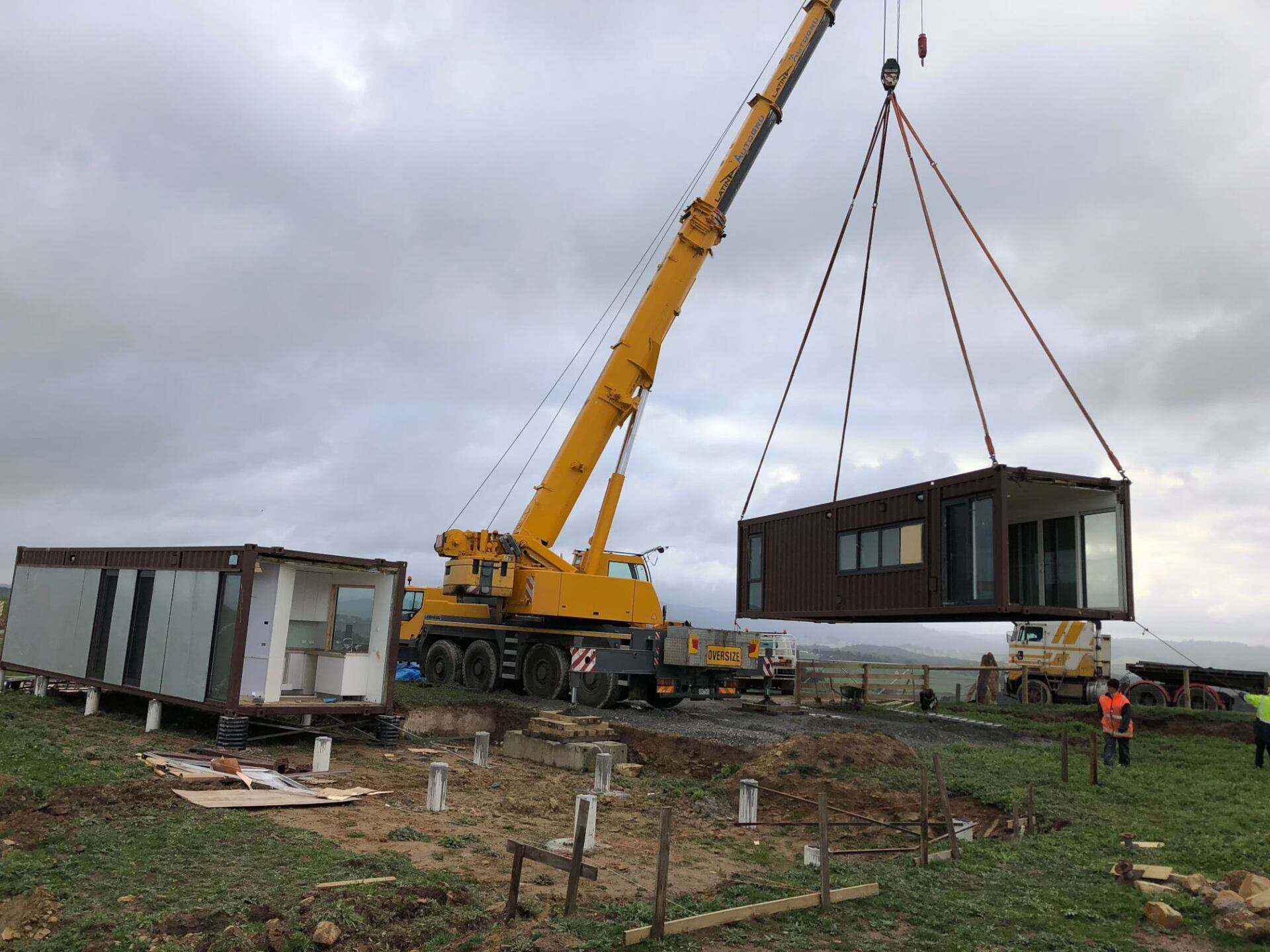A crane lifts a modular building unit; another unit sits on site. Overcast sky, construction area.