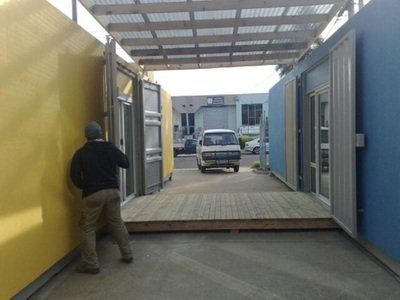 Person by yellow container, looking toward a white van exiting a walkway between blue containers, under a wooden deck.