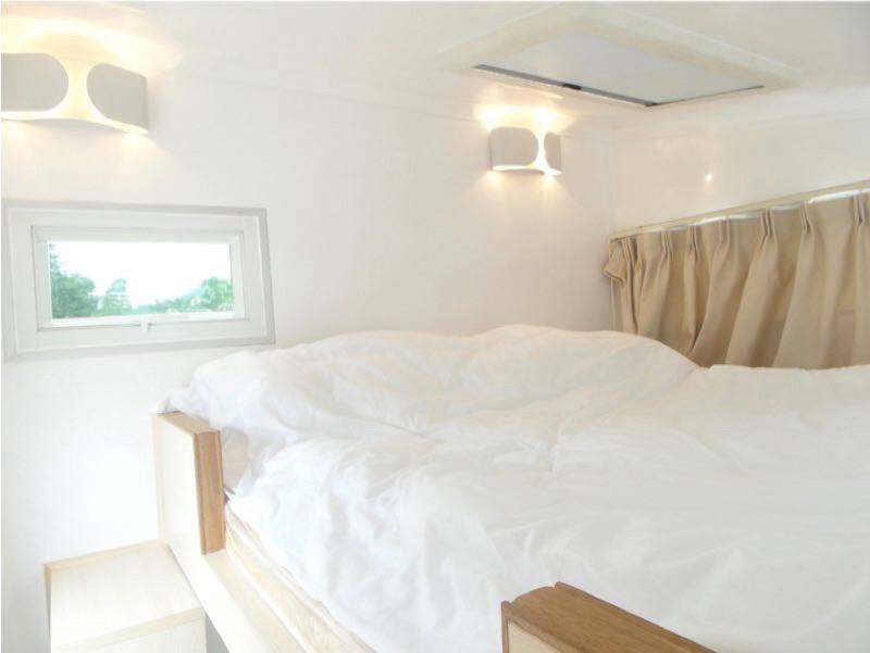 White-walled camper bedroom with a made bed, small window, and wall sconces.