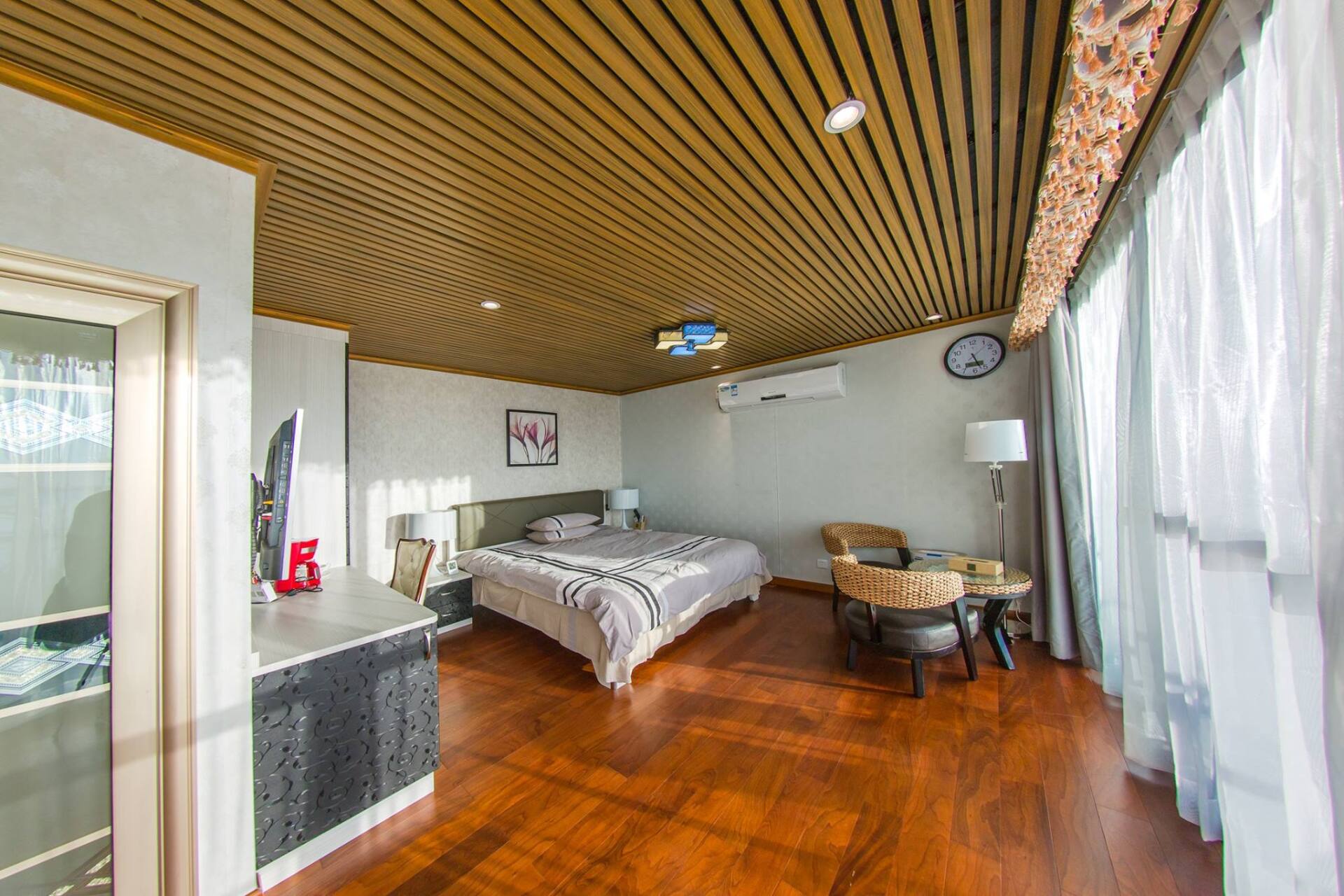 Bedroom with wooden floor and ceiling, bed, sitting area, large window with curtains.