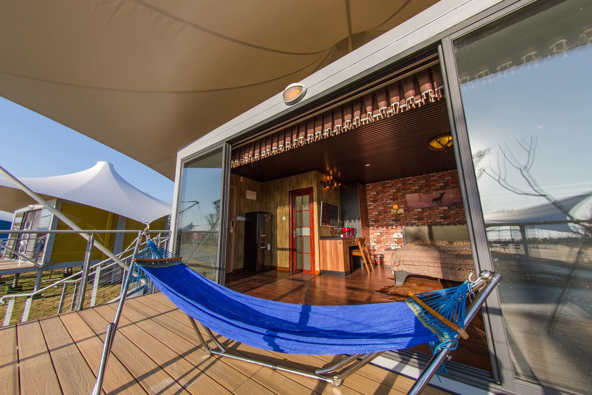 Modern cabin with blue hammock on wooden deck, open glass doors, beige tent roof.