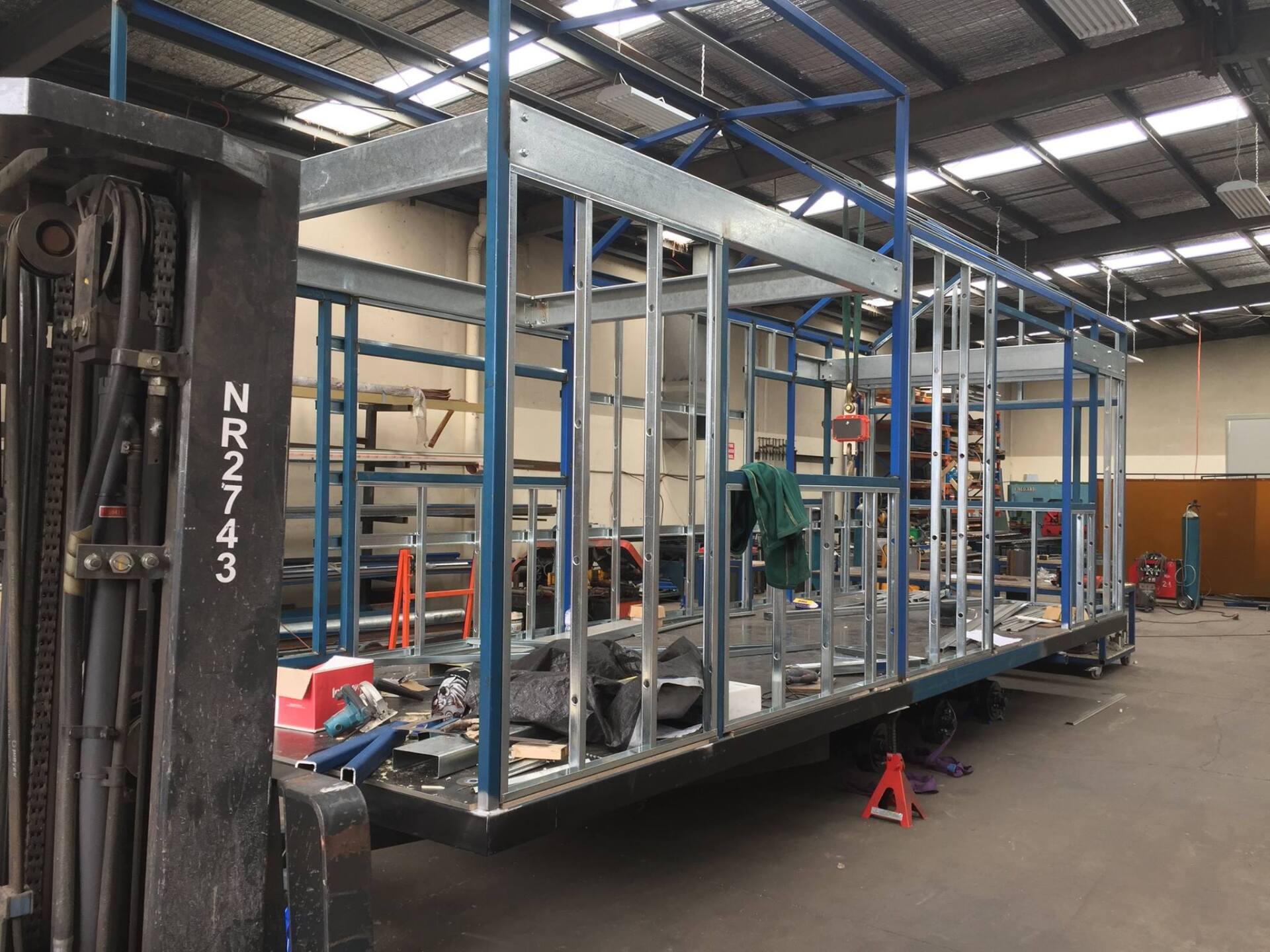 Metal frame of a vehicle being constructed in a workshop. A forklift is to the left.