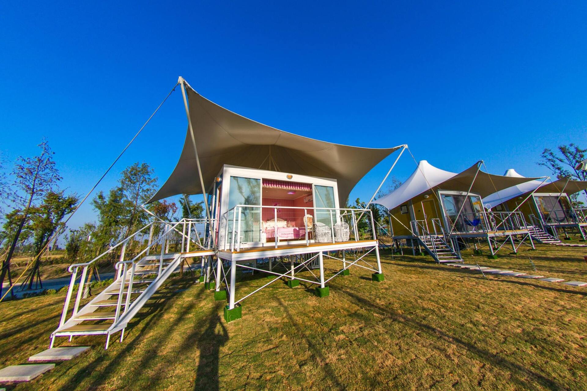 Modern glamping tents with canvas roofs, on a sunny day. White staircases lead to elevated cabins.