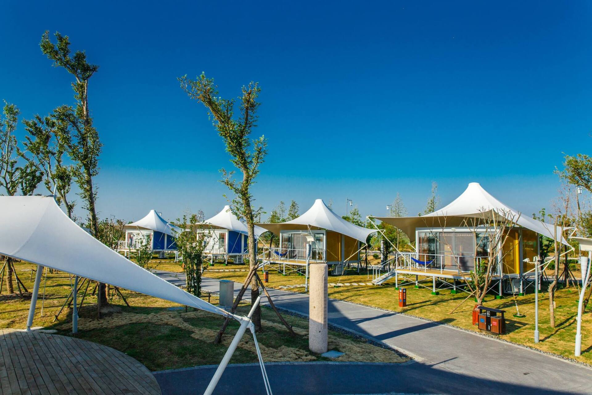 Luxury glamping tents with white canopies and yellow/blue walls sit on grass under a clear blue sky.