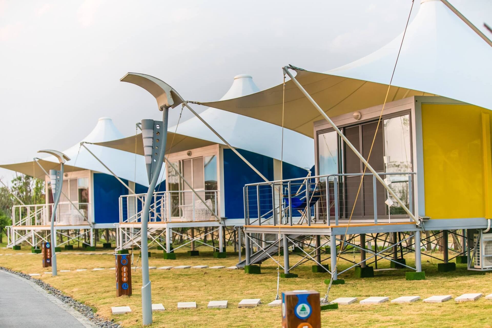 Row of colorful cabins with white tent-like canopies. Each has a porch on a raised platform, set on a grassy area.