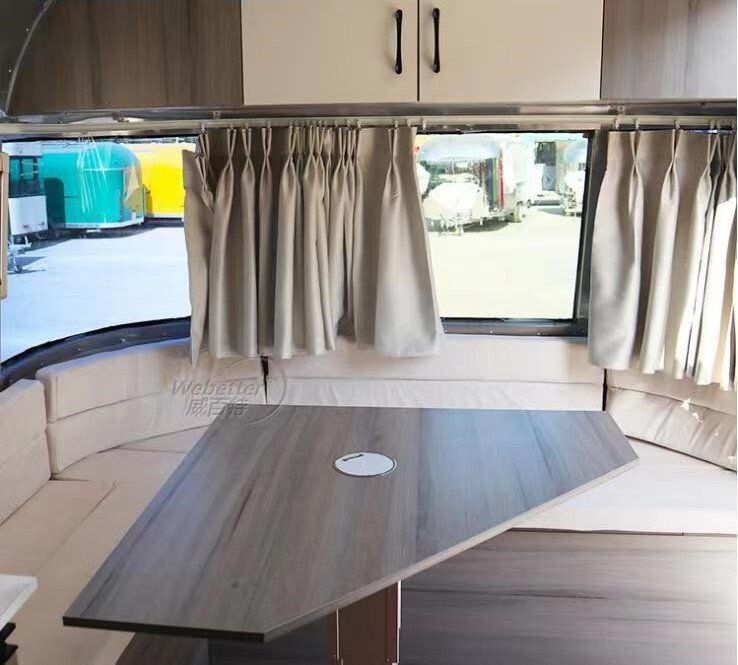 Interior of a trailer with a table, seating, windows, and light-colored curtains.