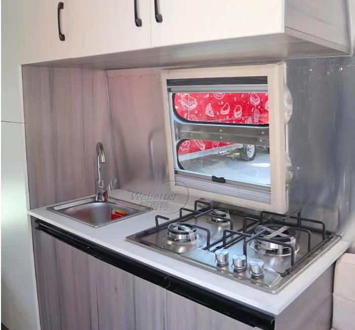 Kitchenette inside a trailer: sink, stove, window, and cabinets. Gray and white surfaces, with a red curtain visible.