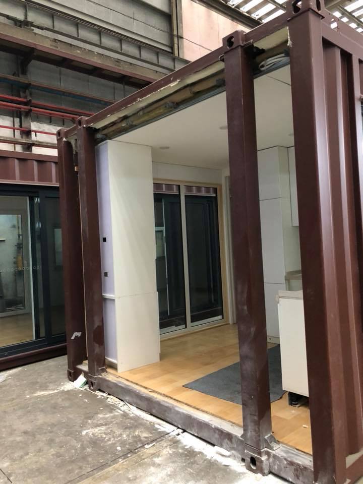 Interior of a modified shipping container, showing a partially built room with a sliding glass door and wooden floor.