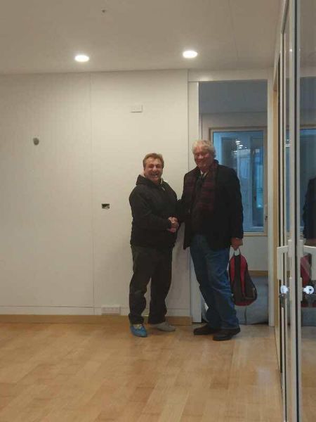 Two men shaking hands in a bare room with a doorway and a mirrored wall.