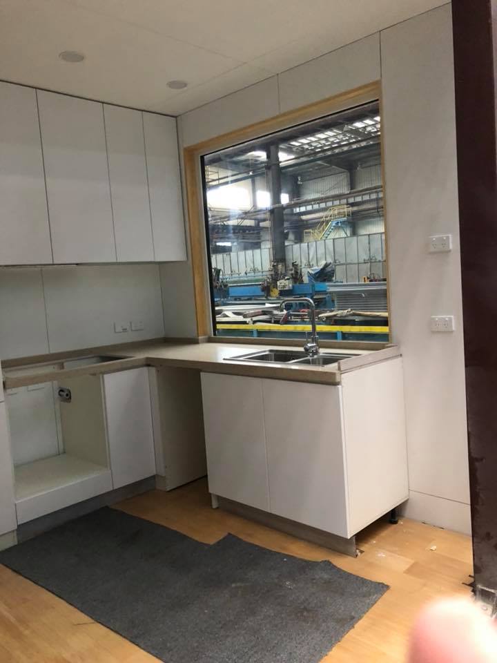 White kitchen cabinets and sink under a window, unfinished, with a view of a factory.