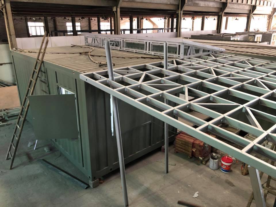 Steel frame construction over a green container. A ladder leans against the side in a warehouse.