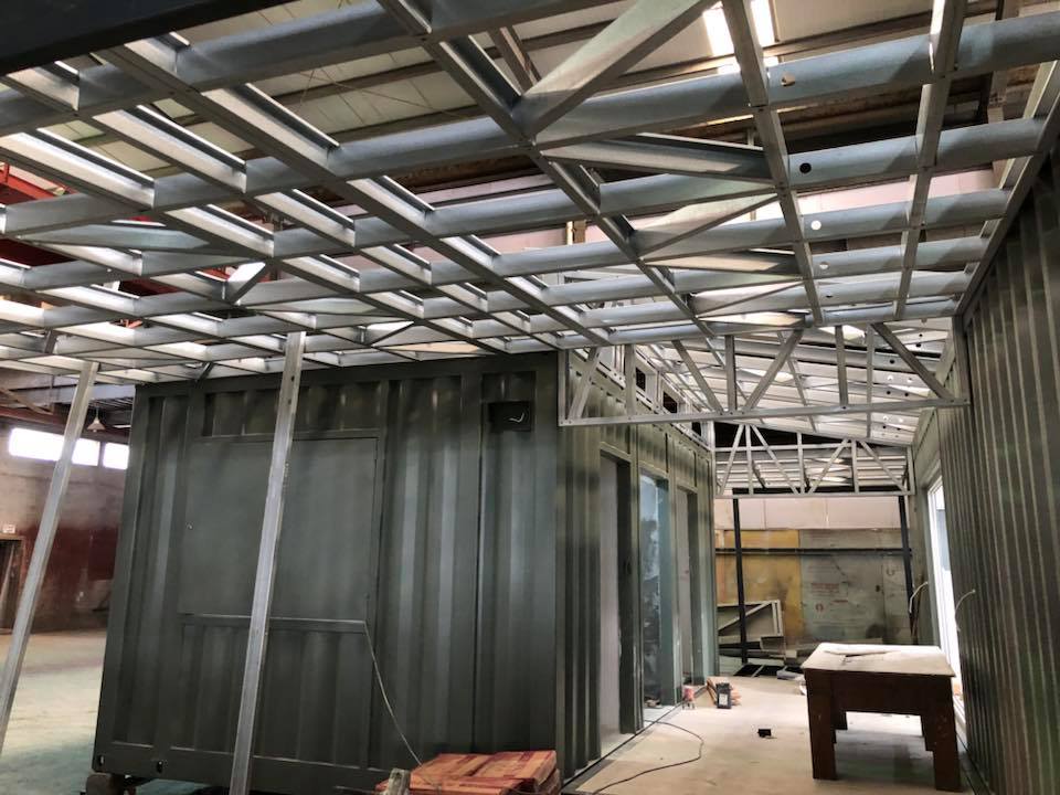 Interior of a building with a metal frame roof and dark container units. Construction in progress.