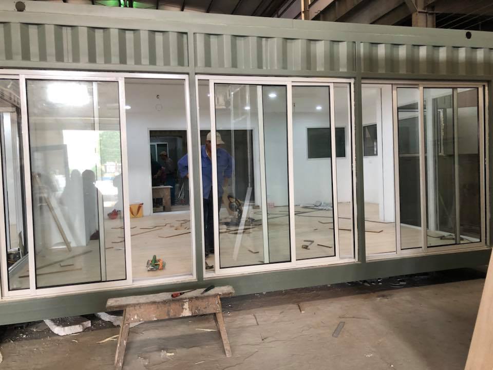 Green shipping container with large glass sliding doors and a partially visible interior, a person reflected in the glass.