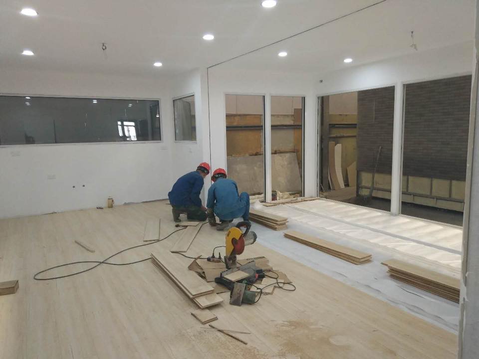 Two workers installing wood flooring in a mostly empty room with construction tools.
