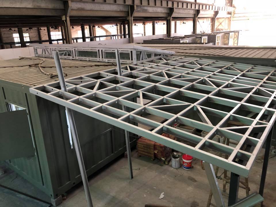 Steel frame being constructed over a green shipping container; indoors with other containers and a metal beam roof.