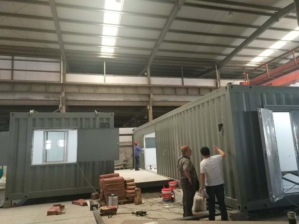 Workers inspecting modular container buildings inside a large industrial facility.