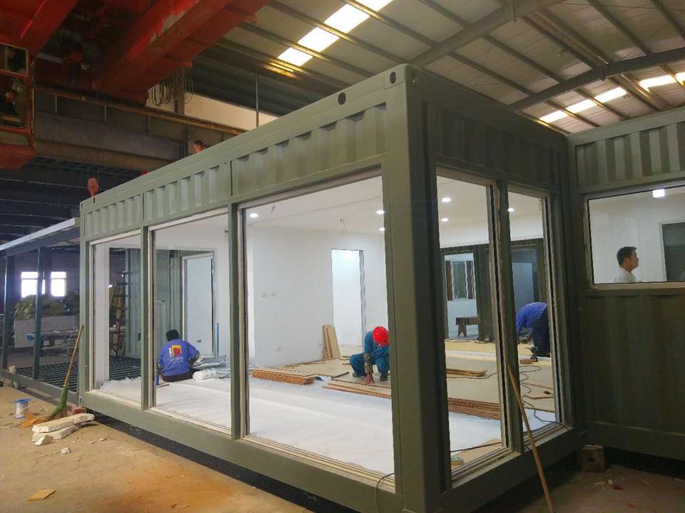 Workers inside a modular building made from shipping containers, with large glass windows and an olive-green exterior, in a factory setting.