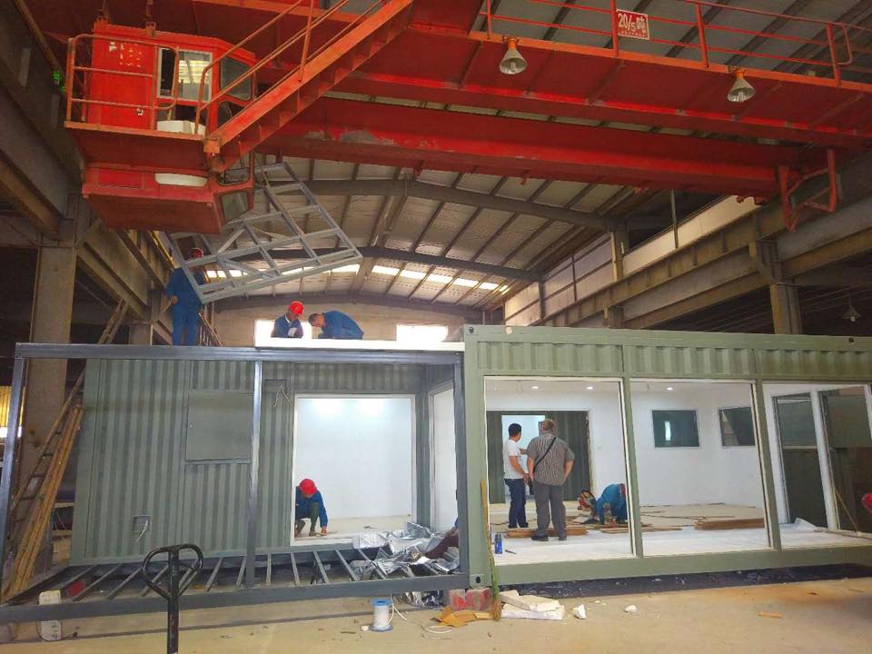Workers assembling modular building units inside a large industrial facility; overhead crane present.