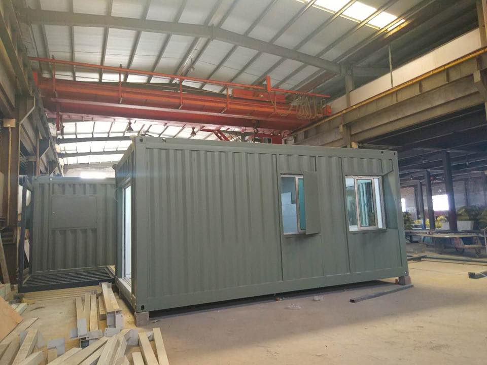 Two olive-green container buildings inside a large industrial facility. One has a window and door, the other a door.