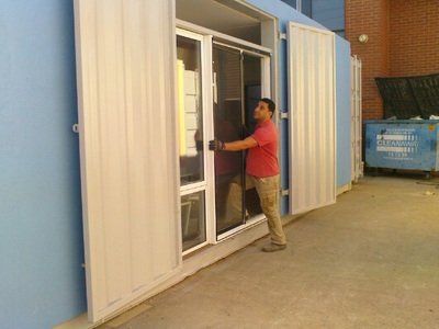 Man opening a sliding metal shutter door next to a glass window. He wears a red shirt and tan pants.
