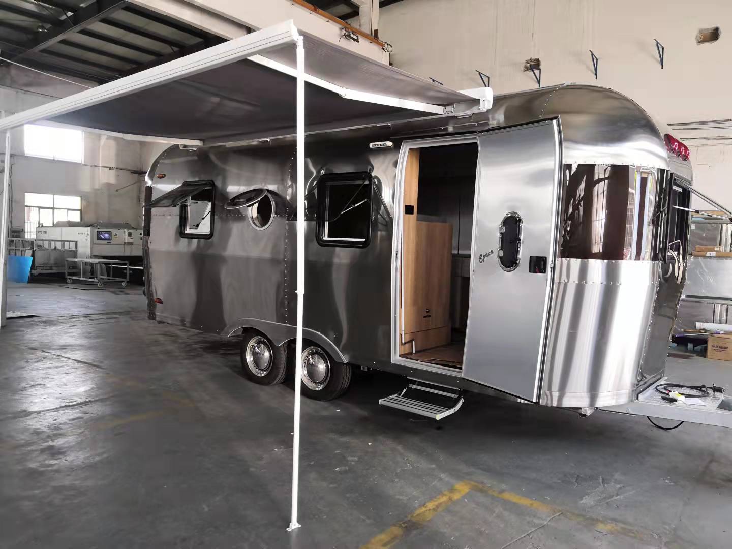Silver, retro-style travel trailer with awning extended, door open, parked inside a building.